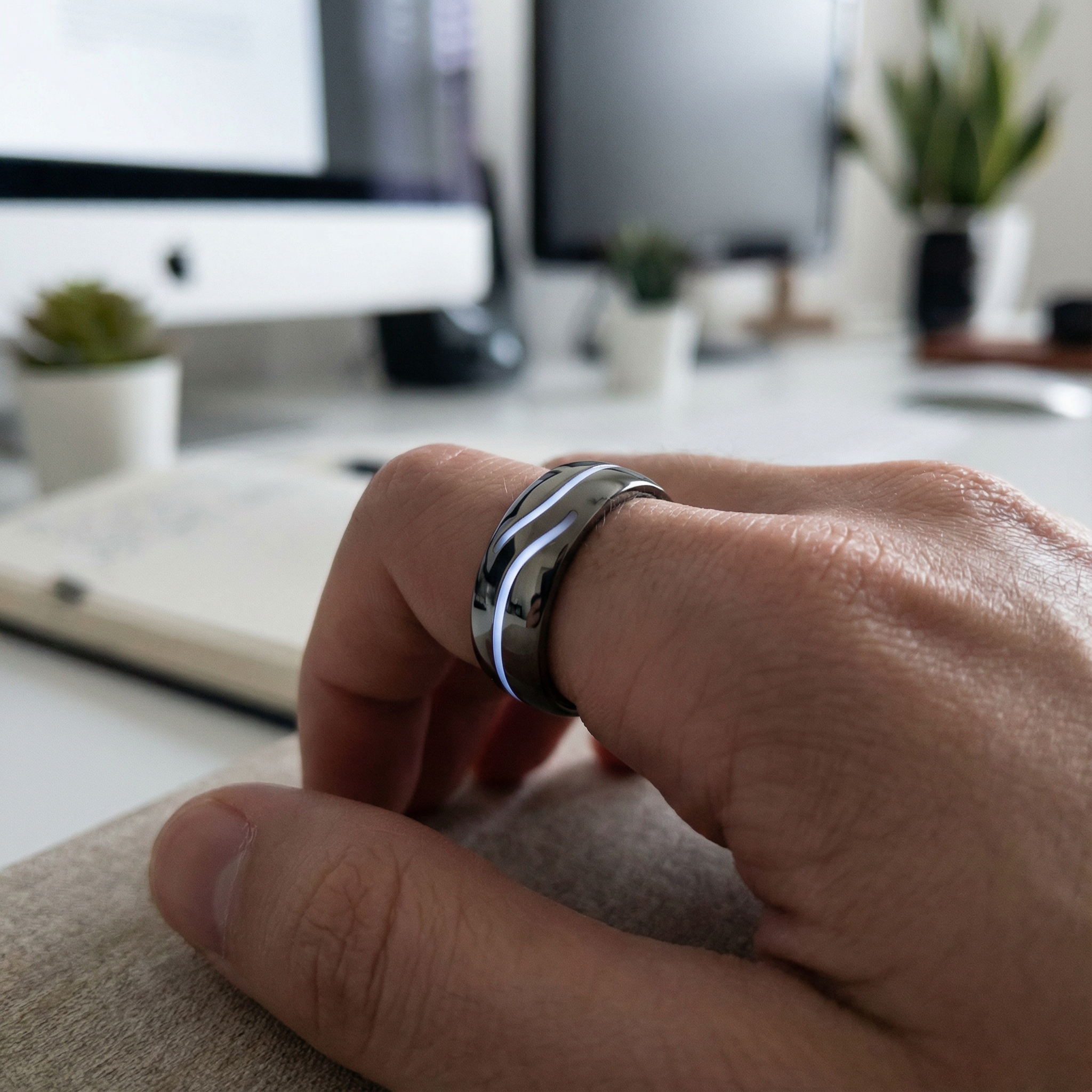 A close-up of a sleek, futuristic ring on an index finger, with subtle glowing elements indicating voice activation. The background is a blurred, modern office or creative space, suggesting idea capture. 1:1 aspect ratio, no visible text.