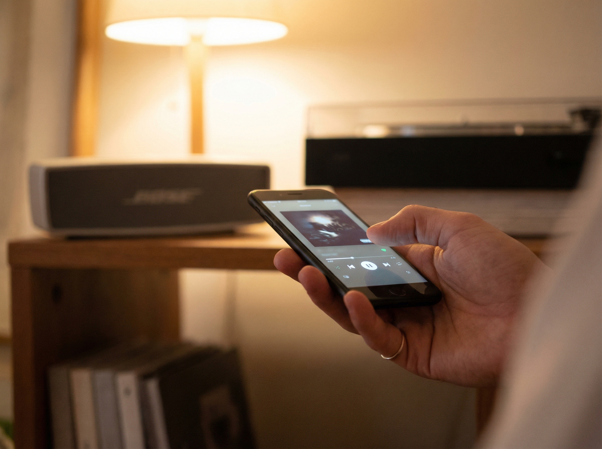 Close up of a person hand holding a smartphone controlling a music application, blurred Bose speaker in the background, soft indoor lighting, realistic lifestyle shot, 4:3 aspect ratio, no text.