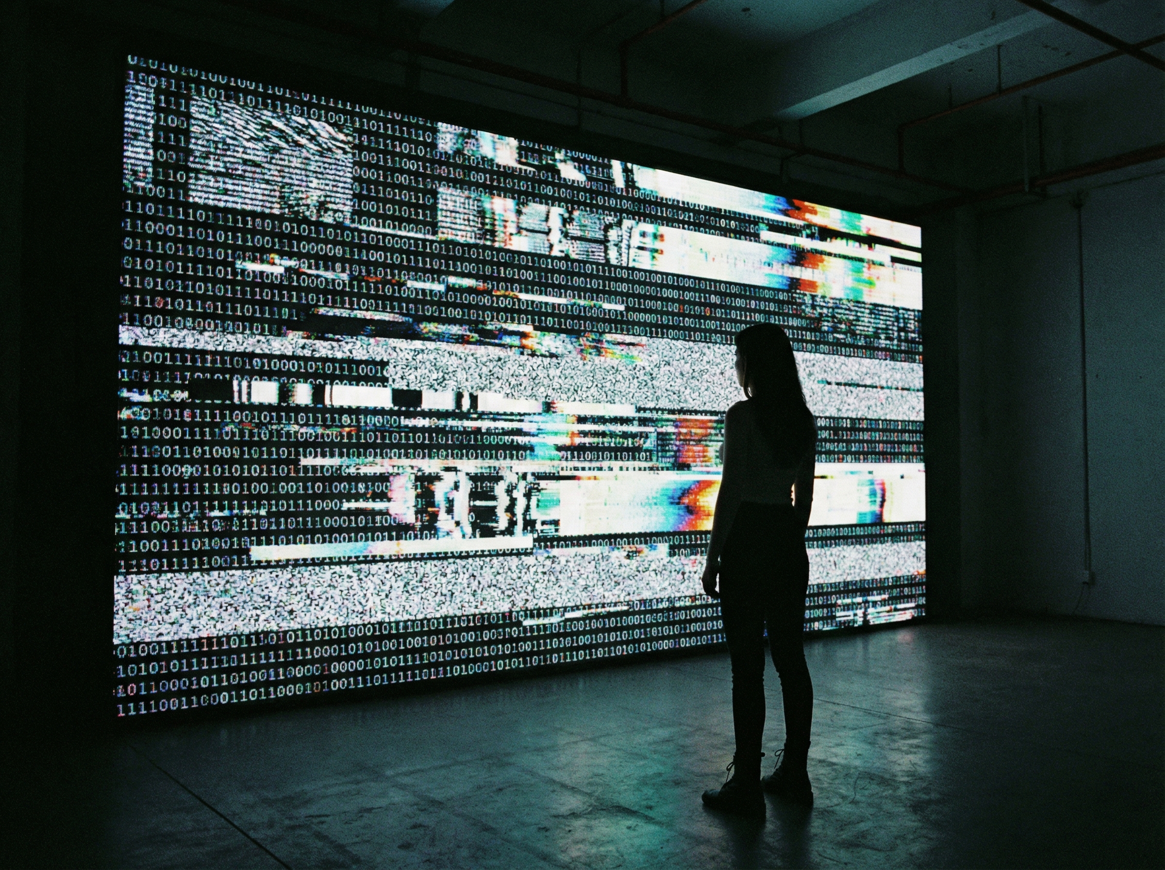 A silhouette of a woman standing in front of a giant digital screen displaying chaotic binary data and distorted patterns, moody lighting, 4:3 aspect ratio, no text