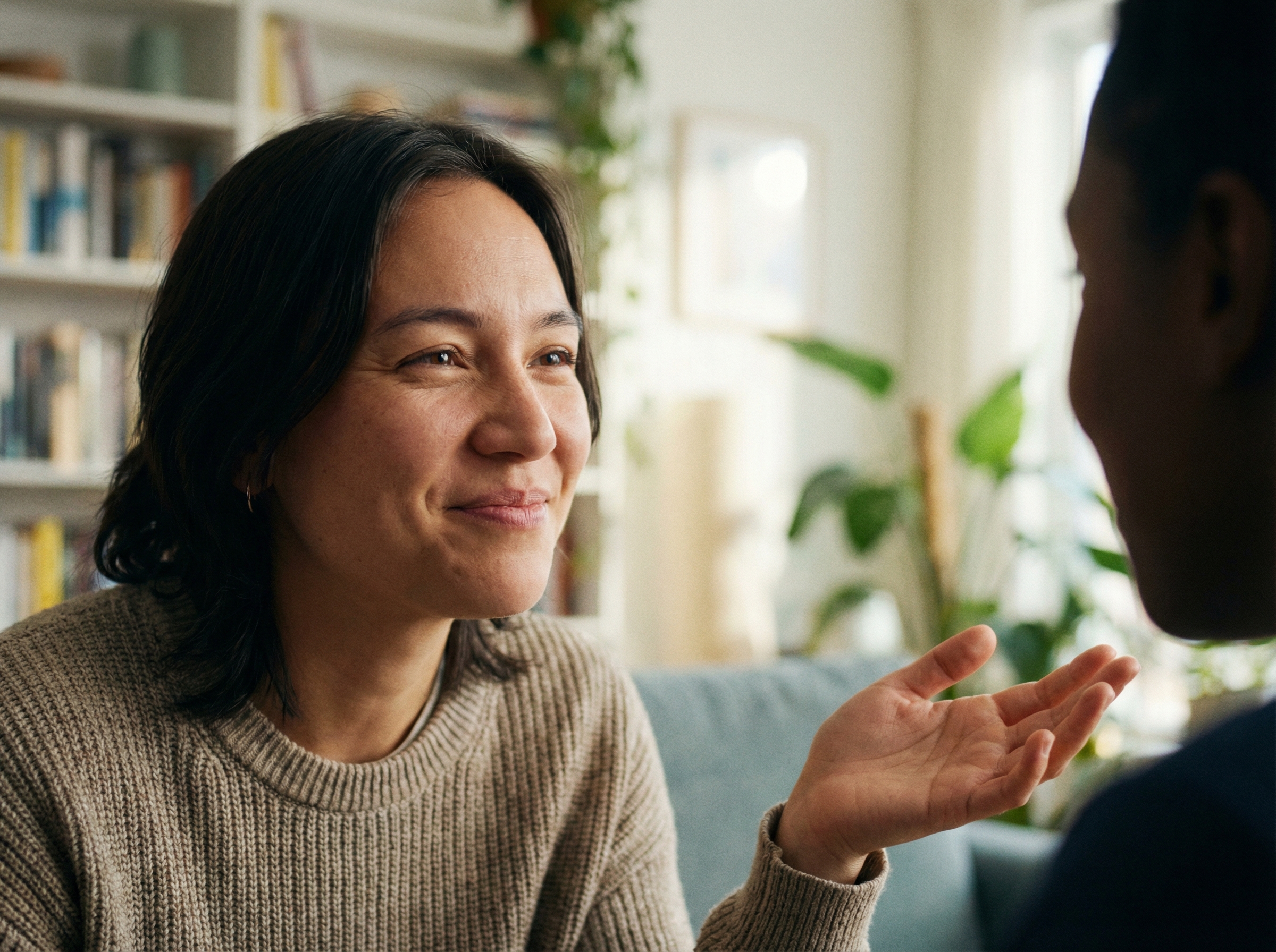 A close-up of a person engaged in a natural, flowing conversation with an unseen AI. The person's expression is relaxed and attentive, as if speaking to a real human. Subtle visual cues like slight head tilt or hand gesture suggest active listening. The background is a soft, out-of-focus home or office environment. No visible text. Aspect ratio 4:3.